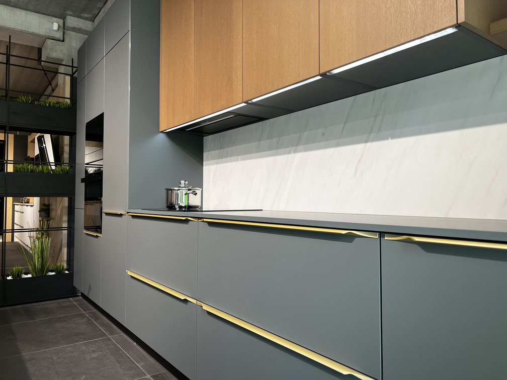 Detail modernej kuchyne NANOTEQ – matné zelene‑sivé skrinky s dlhými mosadznými úchytkami, horné skrinky z dubovej dyhy, biely mramorový obklad a LED podsvietenie; vľavo vstavaná rúra a čierny regál s rastlinami dodávajú industriálny akcent.